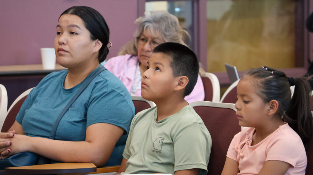 Three people are sitting, one young woman, a male child, and a female child, staring ahead, listening to someone speaking.