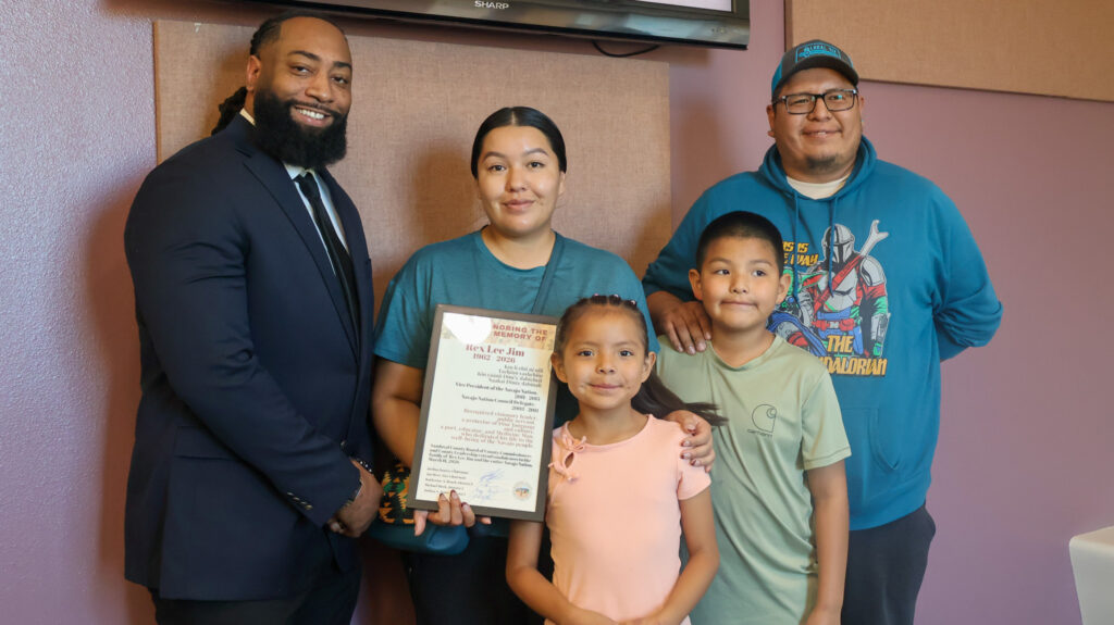 A group of people standing around for a group photo. In the group is Commissioner Joshua Jones standing with Twila Lee, her two children, one girl, one boy, and another male figure. Commissioner Jones is holding up and posing with a piece of paper with the intent of handing the photo to the family.
