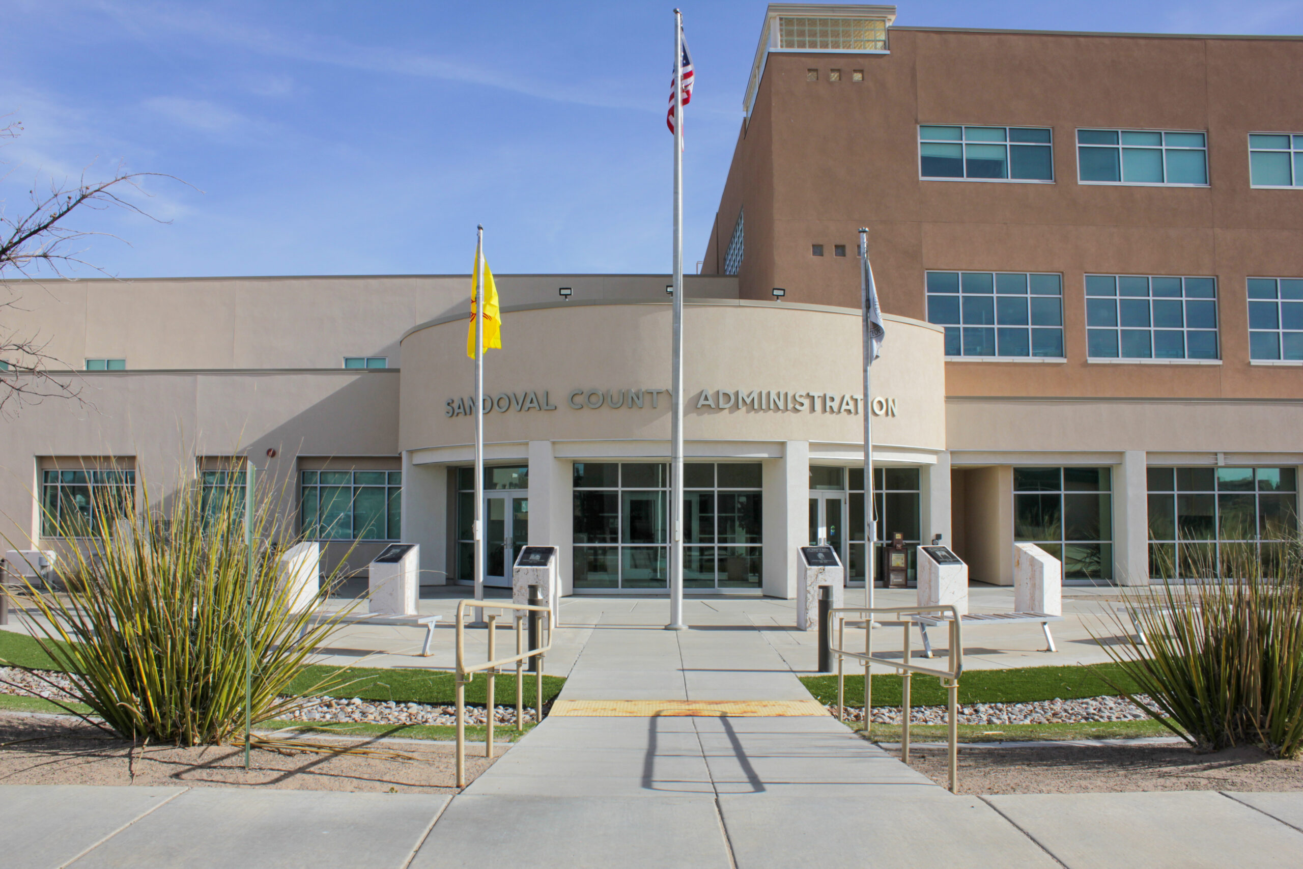 Exterior view of the Sandoval County Administration building with flags and landscaping under a clear blue sky.