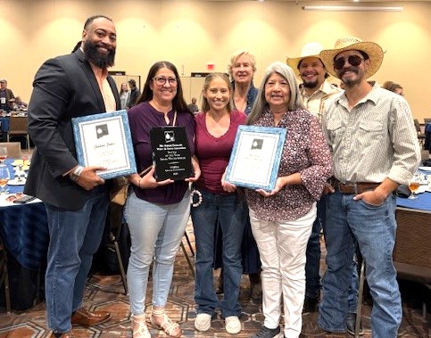 A diverse group of seven individuals stand together in a well-lit room, holding award plaques. The group includes two men wearing cowboy hats. The person on the far left, Commissioner Joshua A. Jones, holds a framed certificate, and the person next to them holds a black plaque. The central figures also hold plaques or certificates, with several individuals wearing jeans and casual attire, while others are dressed more formally in blazers and patterned shirts.