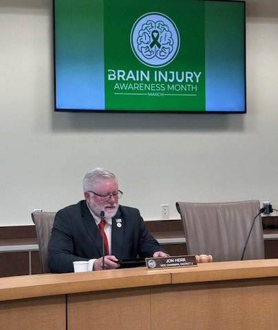A man in a suit and tie sits at a table with a nameplate that reads "Jon Herr, Vice Chairman, District 3," with a large screen behind him displaying "Brain Injury Awareness Month March."
