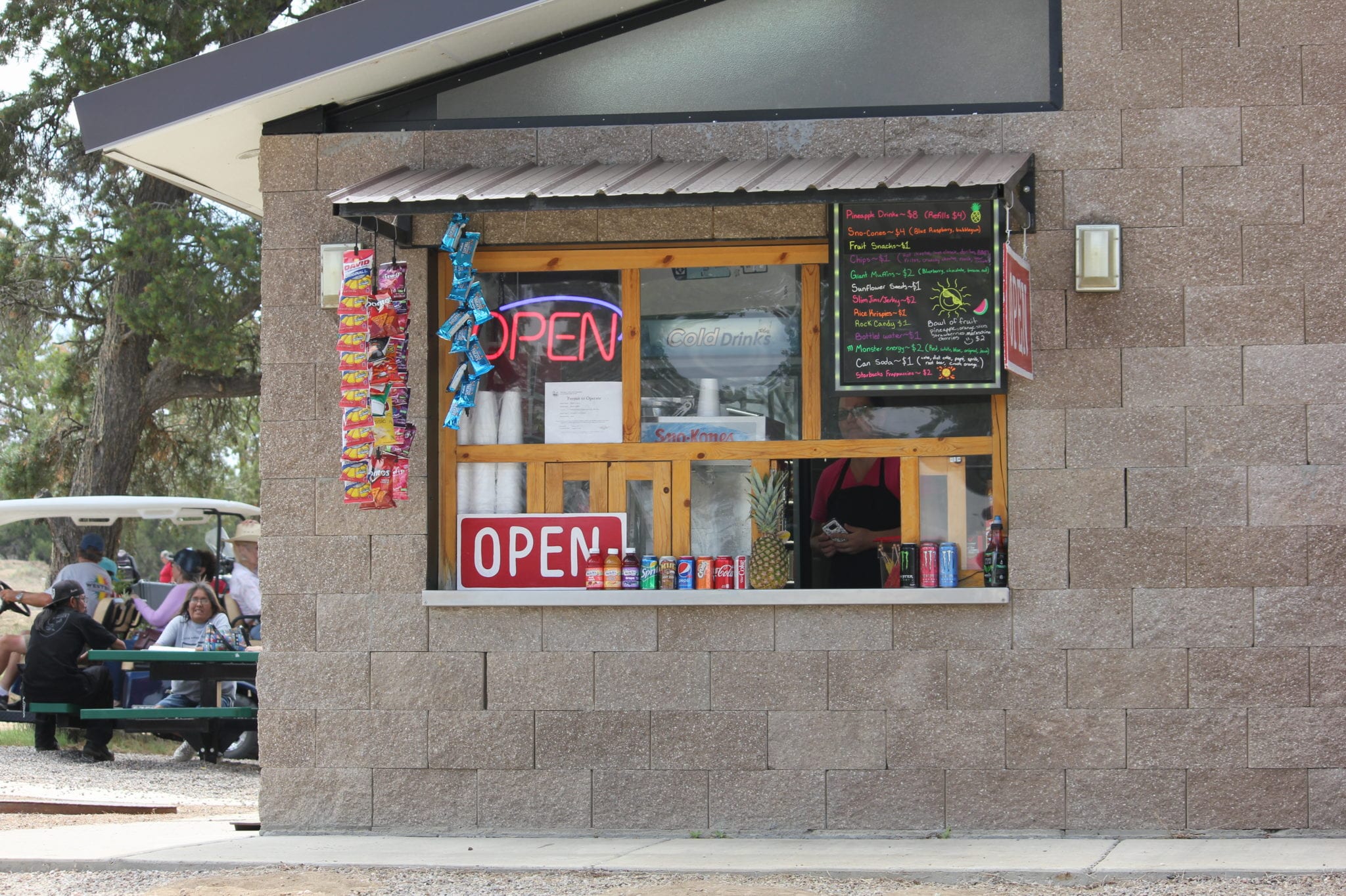 Concession stand window at the Sandoval County Fairgrounds displaying snacks, cold drinks, and a handwritten menu board, with fairgoers seated at a picnic table nearby