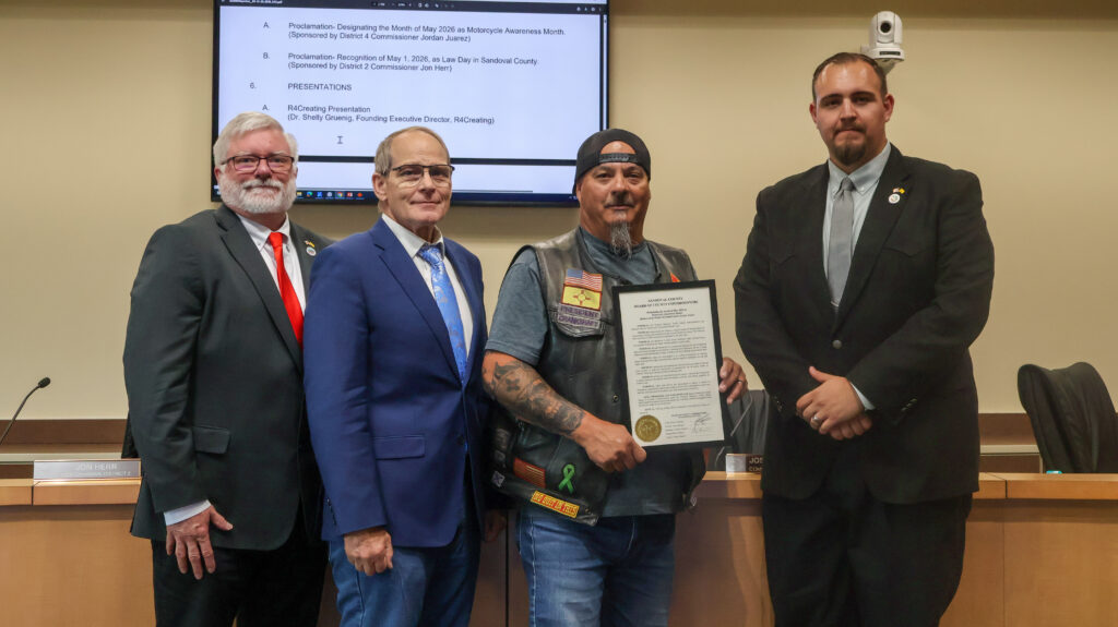 Four men stand formally in what appears to be a government meeting room. The man on the far left wears a dark suit with a red tie and glasses. To his right, a man in a blue suit and patterned tie smiles broadly. The central figure, wearing a black biker vest over a t-shirt, a black baseball cap, and jeans, holds up a framed certificate. The man on the far right is dressed in a black suit and grey tie, standing attentively with his arms crossed. A projector screen with text is visible behind them.