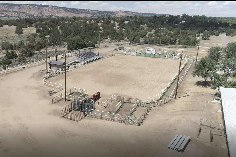 Aerial view of the Sandoval County Fairgrounds rodeo arena showing the dirt competition floor, white pipe-rail fencing, bucking chutes, bleacher seating, and surrounding high desert landscape