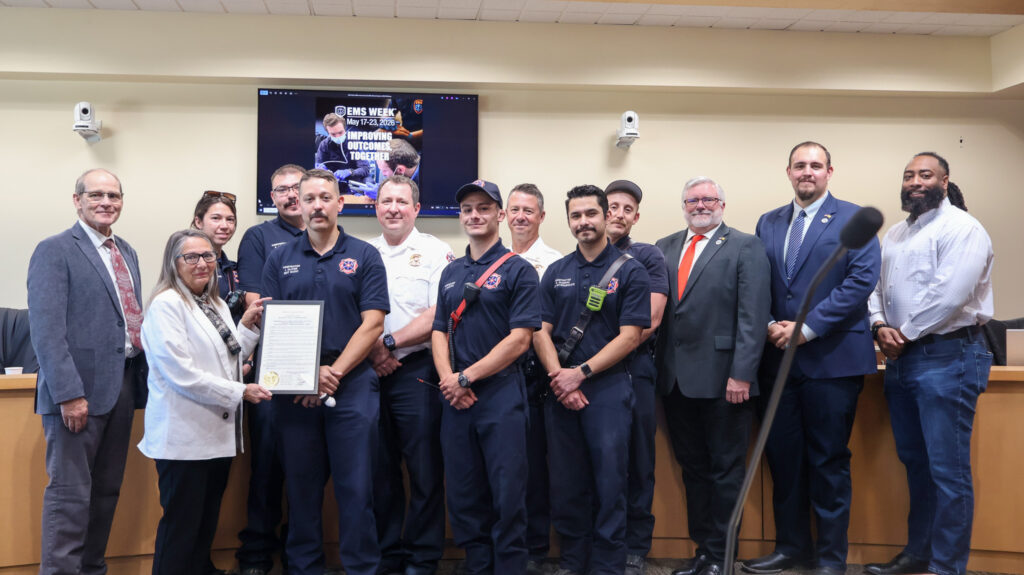 A group of emergency medical service personnel and civilians stand together for a photo, with a woman holding a framed certificate, in front of a screen displaying "EMS WEEK" and a microphone stand.