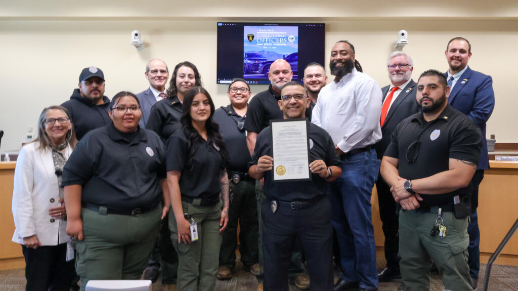 Group of correctional officers and officials pose for a photo in front of a screen displaying "National Correctional Officers Week." One officer in the center holds up a framed certificate.