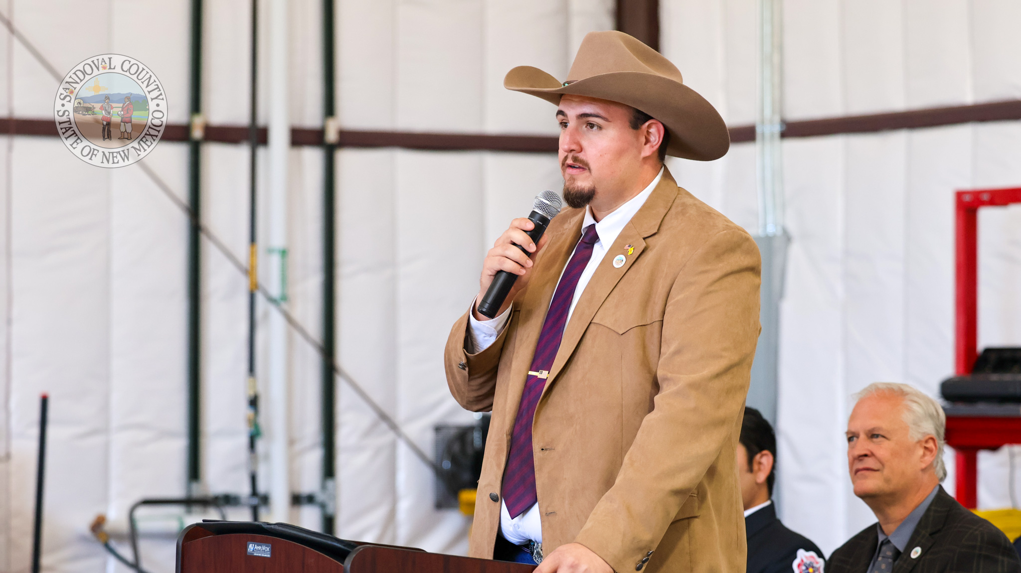 IMG_0401 A man in a tan cowboy hat and blazer speaks into a microphone at a podium, with a county seal visible on the wall behind him.