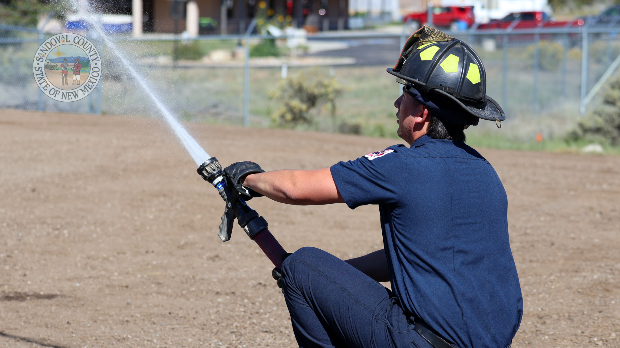 IMG_0439 A firefighter wearing a dark blue uniform and a black helmet with yellow reflective markings sprays water from a hose.